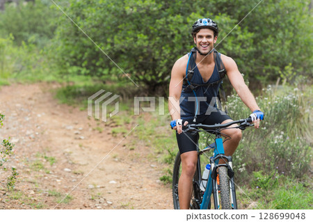Pedaling man riding mountain bike on narrow trail in woods, with hydration pack, helmet, copy space Pedaling man riding mountain bike on narrow trail in woods, with hydration pack, helmet, copy space 128699048