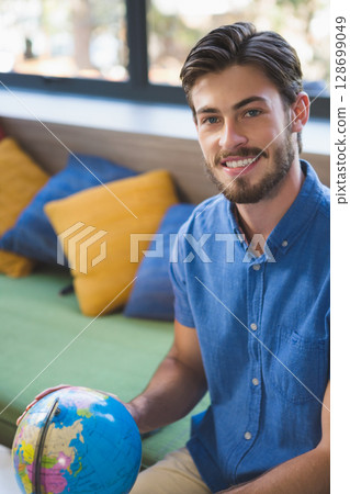 Smiling man sitting on green upholstered bench in modern lounge, holding small world globe Smiling man sitting on green upholstered bench in modern lounge, holding small world globe 128699049