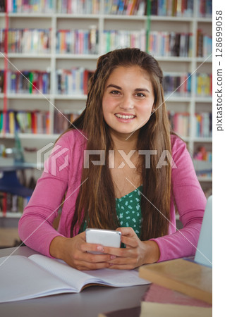 Smiling teenage girl holding smartphone at library table, with notebook, laptop and textbooks 128699058