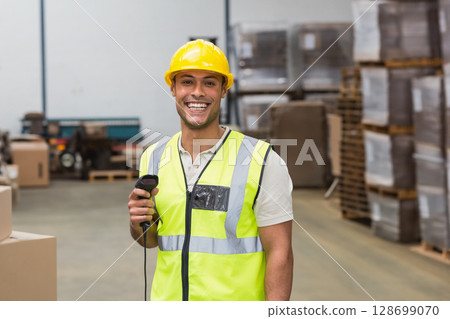 Smiling male warehouse worker holding barcode scanner in busy distribution center with pallets 128699070