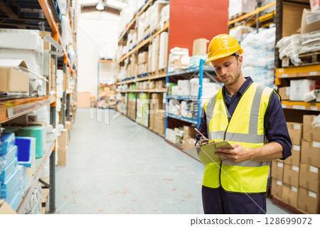 Male warehouse worker inspecting inventory in warehouse aisle, with clipboard and pen, copy space 128699072