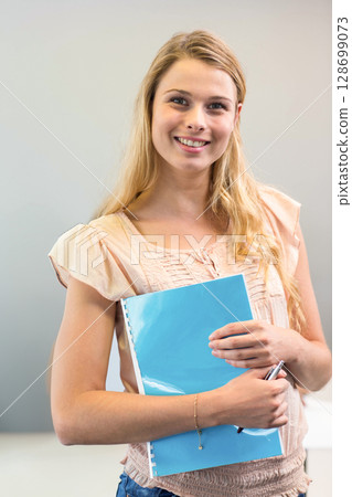Smiling teenage girl holding blue binder and pen against light gray wall, wearing gold bracelet Smiling teenage girl holding blue binder and pen against light gray wall, wearing gold bracelet 128699073