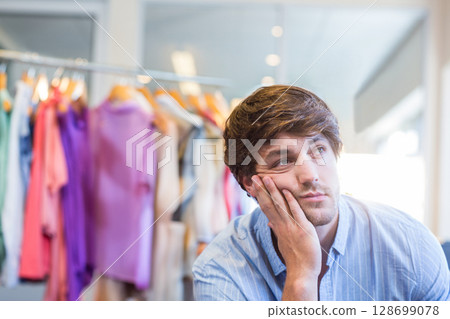 Leaning male shopper touching chin at clothing store, with pastel shirts on metal rack, copy space 128699078