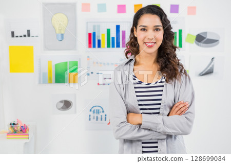 Standing woman crossing arms and smiling in office, with chart wall and sticky notes, copy space 128699084