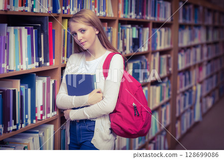Leaning teenage girl holding blue book in library aisle, with red backpack and wooden shelves 128699086