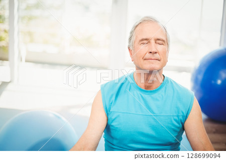 Meditating Senior man sitting cross-legged on floor in fitness studio, with blue stability balls 128699094