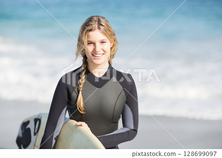 Smiling woman holding surfboard and wearing wetsuit on sandy beach, under clear blue sky Smiling woman holding surfboard and wearing wetsuit on sandy beach, under clear blue sky 128699097