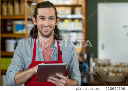 Smiling man standing behind store counter, wearing red apron and holding tablet near wicker baskets 128699106