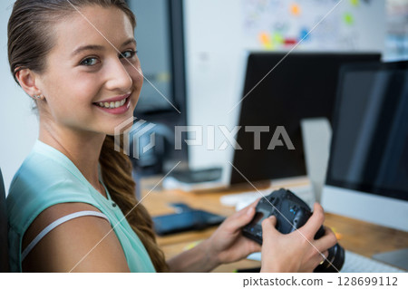 Smiling woman holding DSLR camera at wooden desk in office, with computer monitors, copy space Smiling woman holding DSLR camera at wooden desk in office, with computer monitors, copy space 128699112