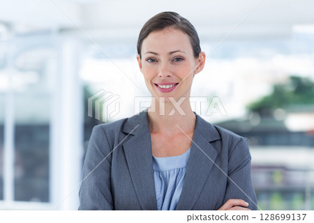 Smiling woman standing with arms crossed in modern office, with large windows and urban view 128699137