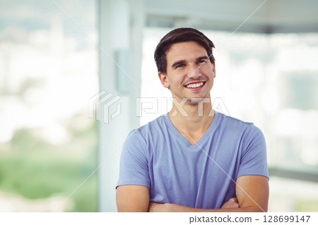 Smiling male in mid-20s standing with arms crossed in modern interior, featuring blurred cityscape 128699147