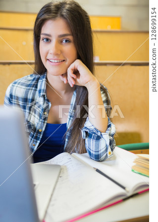 Working female student taking notes on notebook at university lecture hall desk, using laptop 128699154