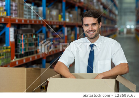 Smiling man handling large cardboard box in warehouse aisle, with metal shelving racks 128699163