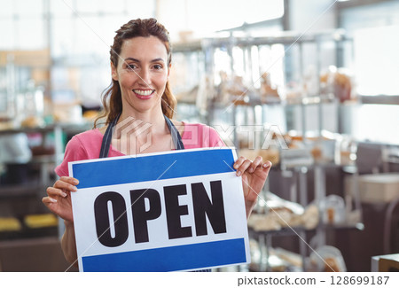 Smiling woman baker holding OPEN sign inside bakery, with metal racks and pastries Smiling woman baker holding OPEN sign inside bakery, with metal racks and pastries 128699187