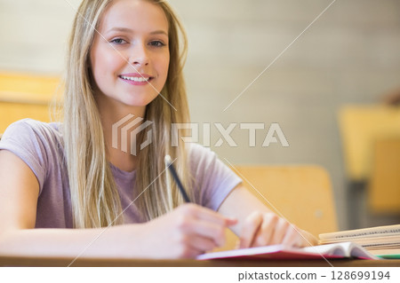 Smiling teenage girl writing in classroom at wooden school desk, with open notebook and pencil 128699194