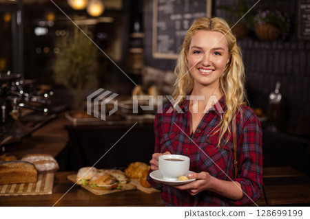 Smiling woman holding cup and saucer with cookie at coffee shop counter, baked goods, copy space Smiling woman holding cup and saucer with cookie at coffee shop counter, baked goods, copy space 128699199