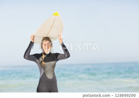 Smiling woman wearing wetsuit standing at beach shore, holding white surfboard overhead 128699200
