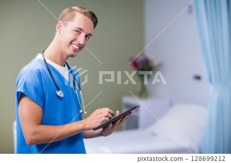 Smiling male doctor holding tablet and stethoscope beside hospital bed in patient room, copy space 128699212