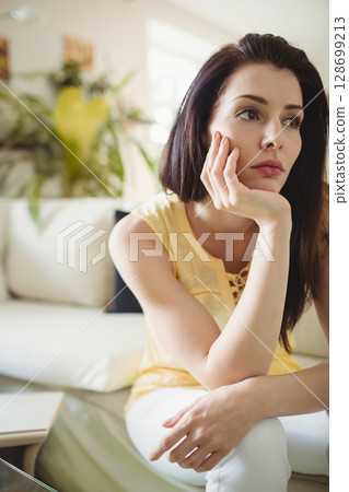 Leaning woman resting chin on hand in modern living room, with laptop and potted plant 128699213