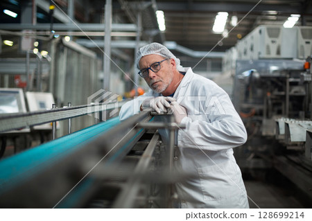 Senior man wearing lab coat and hairnet leaning on conveyor belt railing in factory, with machinery Senior man wearing lab coat and hairnet leaning on conveyor belt railing in factory, with machinery 128699214