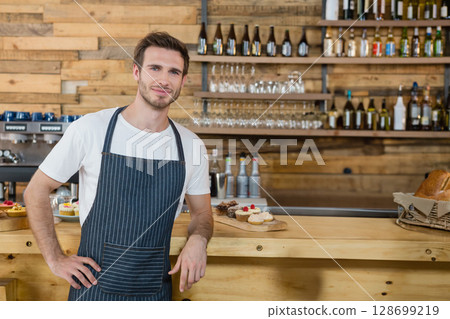 Smiling male barista leaning on rustic wooden counter in cafe, showcasing assorted pastries, bread Smiling male barista leaning on rustic wooden counter in cafe, showcasing assorted pastries, bread 128699219
