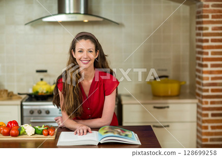 Smiling woman referencing open cookbook at home kitchen countertop, with cutting board and veggies Smiling woman referencing open cookbook at home kitchen countertop, with cutting board and veggies 128699258