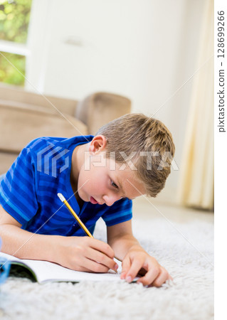 Boy lying on white carpet in bright living room, writing in open notebook with yellow pencil Boy lying on white carpet in bright living room, writing in open notebook with yellow pencil 128699266