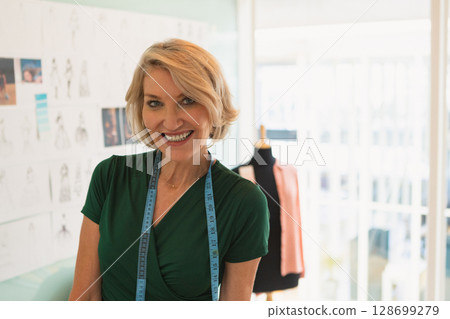 Senior woman standing in design studio, wearing blue tape and showing pinned sketches, coral jacket Senior woman standing in design studio, wearing blue tape and showing pinned sketches, coral jacket 128699279
