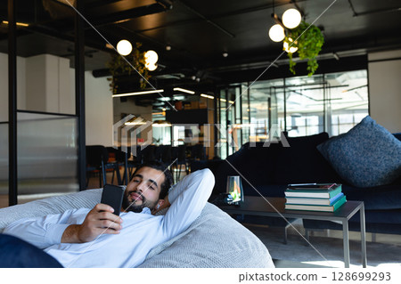 Lounging man reclining on gray beanbag chair in coworking lounge, holding smartphone 128699293