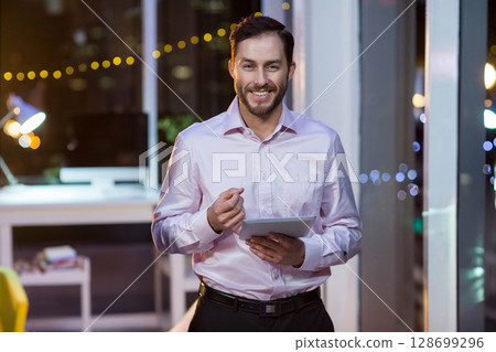 Holding tablet and gesturing, male professional working late in office, with desk lamp and monitor 128699296
