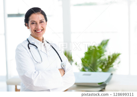 Smiling senior woman doctor crossing arms while standing in clinic, with stethoscope, copy space Smiling senior woman doctor crossing arms while standing in clinic, with stethoscope, copy space 128699311