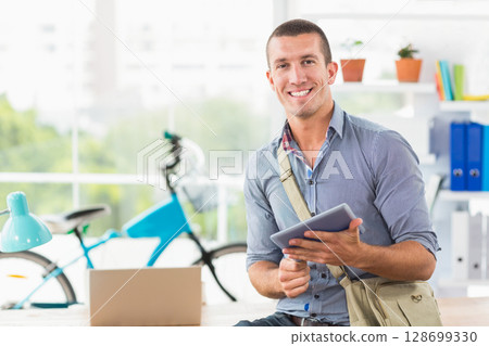 Smiling man holding tablet at modern office desk, with box, desk lamp and bicycle, copy space 128699330