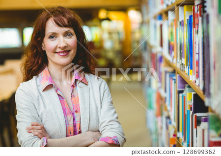 Standing woman librarian folding arms in public library aisle, with bookshelf loaded with books 128699362