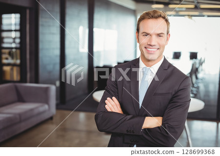 Standing middle-aged man wearing black suit crossing arms in open-plan office, glass partitions Standing middle-aged man wearing black suit crossing arms in open-plan office, glass partitions 128699368