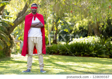Standing African American man posing in sunlit backyard, with red superhero cape and mask Standing African American man posing in sunlit backyard, with red superhero cape and mask 128699387