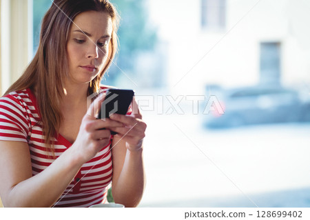Browsing woman sitting by large window indoors, using black smartphone under soft natural light Browsing woman sitting by large window indoors, using black smartphone under soft natural light 128699402