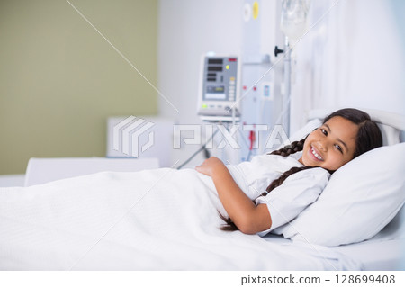 Smiling female child patient resting on pediatric ward bed with infusion pump tubing, copy space 128699408