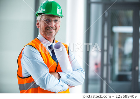 Inspecting man wearing hard hat and safety vest inside lobby, holding blueprints, copy space 128699409