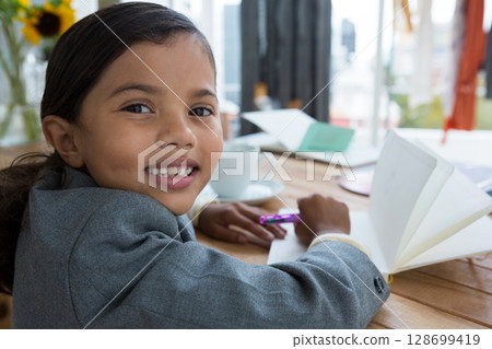 Writing girl child using purple pen over open notebook at table near window, with sunflower vase Writing girl child using purple pen over open notebook at table near window, with sunflower vase 128699419