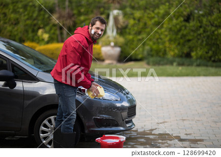 Scrubbing man cleaning hood of hatchback on driveway, with yellow sponge and red bucket, copy space 128699420