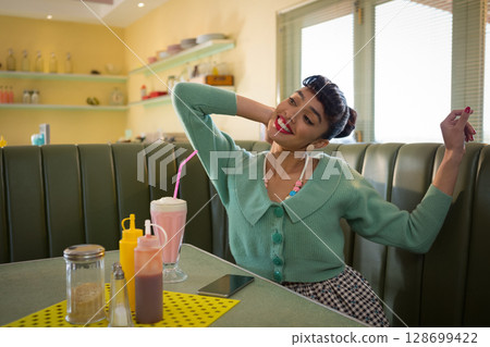 Smiling woman stretching arms behind head at diner booth, with milkshake and smartphone, copy space 128699422