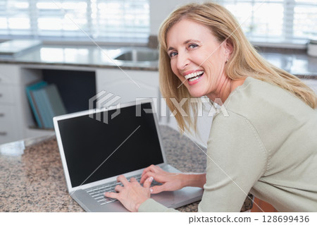 Standing senior woman typing on laptop at kitchen countertop with stainless steel sink, copy space Standing senior woman typing on laptop at kitchen countertop with stainless steel sink, copy space 128699436