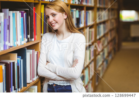 Smiling teenage girl leaning against bookshelf in library aisle, with colorful books, copy space Smiling teenage girl leaning against bookshelf in library aisle, with colorful books, copy space 128699447