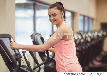 Exercising woman standing on elliptical in fitness center, with machine handles and mirror 128699448