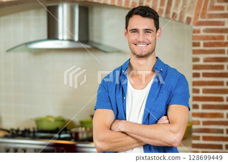 Smiling man standing with arms crossed in modern kitchen, featuring steel range hood and green pots 128699449
