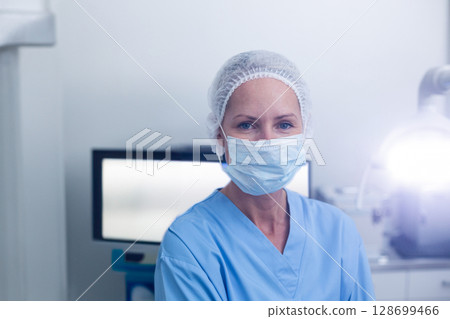 Standing female medical worker wearing scrubs and mask in operating room, with monitor and light 128699466