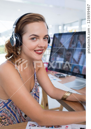 Smiling woman wearing headphones working at desk in office, with computer monitor, copy space Smiling woman wearing headphones working at desk in office, with computer monitor, copy space 128699467