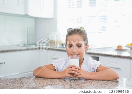Smiling child girl sitting at granite countertop in white kitchen, holding clear glass of milk Smiling child girl sitting at granite countertop in white kitchen, holding clear glass of milk 128699486