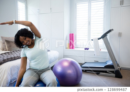 Stretching mid-adult African American woman sitting on ball in home gym, with yoga mats, treadmill 128699487