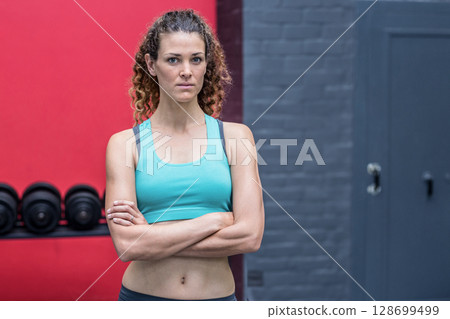 Standing woman crossing arms at gym weight area, with loaded dumbbell rack against red wall 128699499